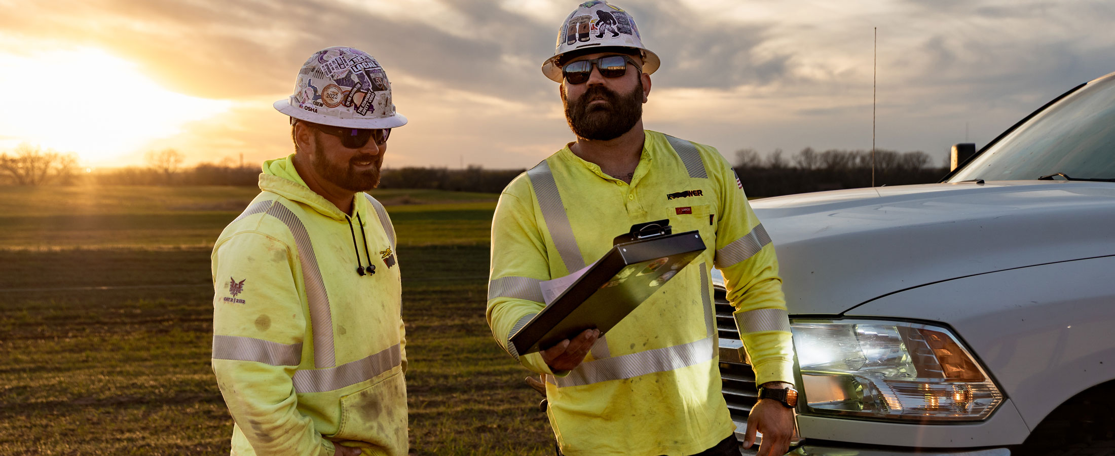 Two electrical linemen, Dylan Edgmon and Kevin Walker, in high-visibility jackets and hard hats stand beside a work truck at sunset, one holding a clipboard as they review a task.
